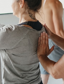 כף יד מנחה מונחת על הגב העליון של מתאמנת בחולצה אפורה עם קעקוע בעורף - תיקון מנח ויציבה בשיעור.